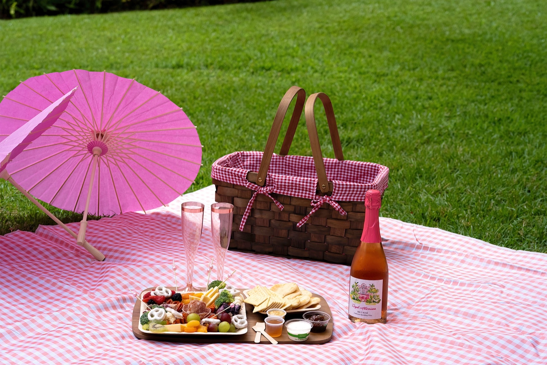 芝生の上に広げたピンクのギンガムチェックのレジャーシートの上で、ピクニックバスケットとスパークリングワイン、軽食が並ぶピクニックセット