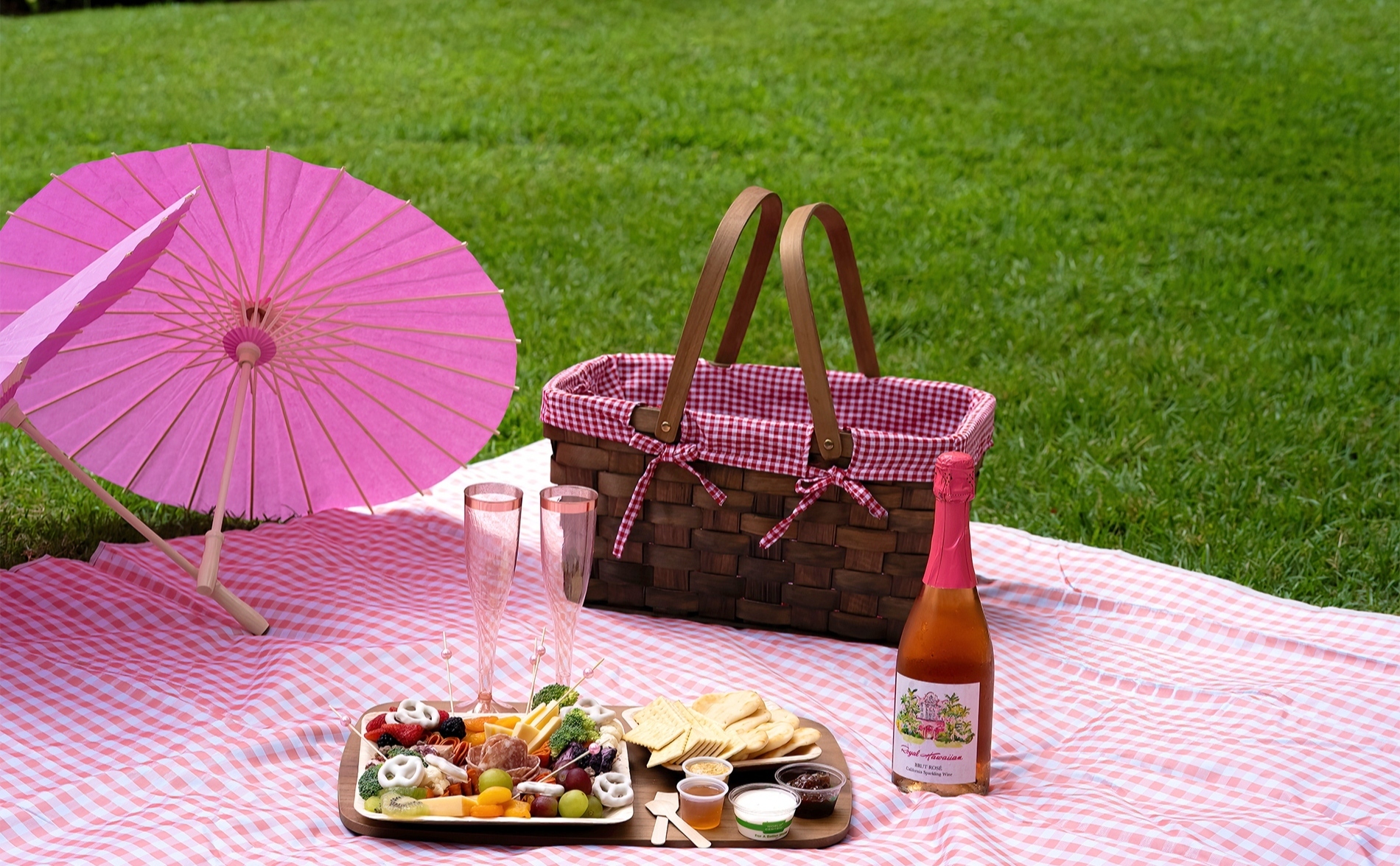 芝生の上に広げたピンクのギンガムチェックのレジャーシートの上で、ピクニックバスケットとスパークリングワイン、軽食が並ぶピクニックセット